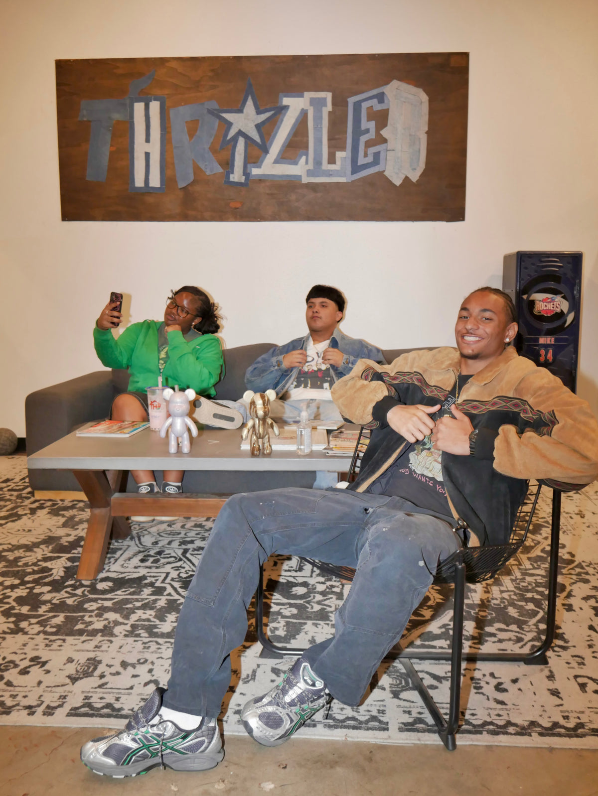 Three people posing in a modern living room setting with a large wooden 'THRILLER' sign on the wall behind them. The person in the foreground is wearing a tan jacket, black shirt, and gray sneakers, sitting in a black chair.