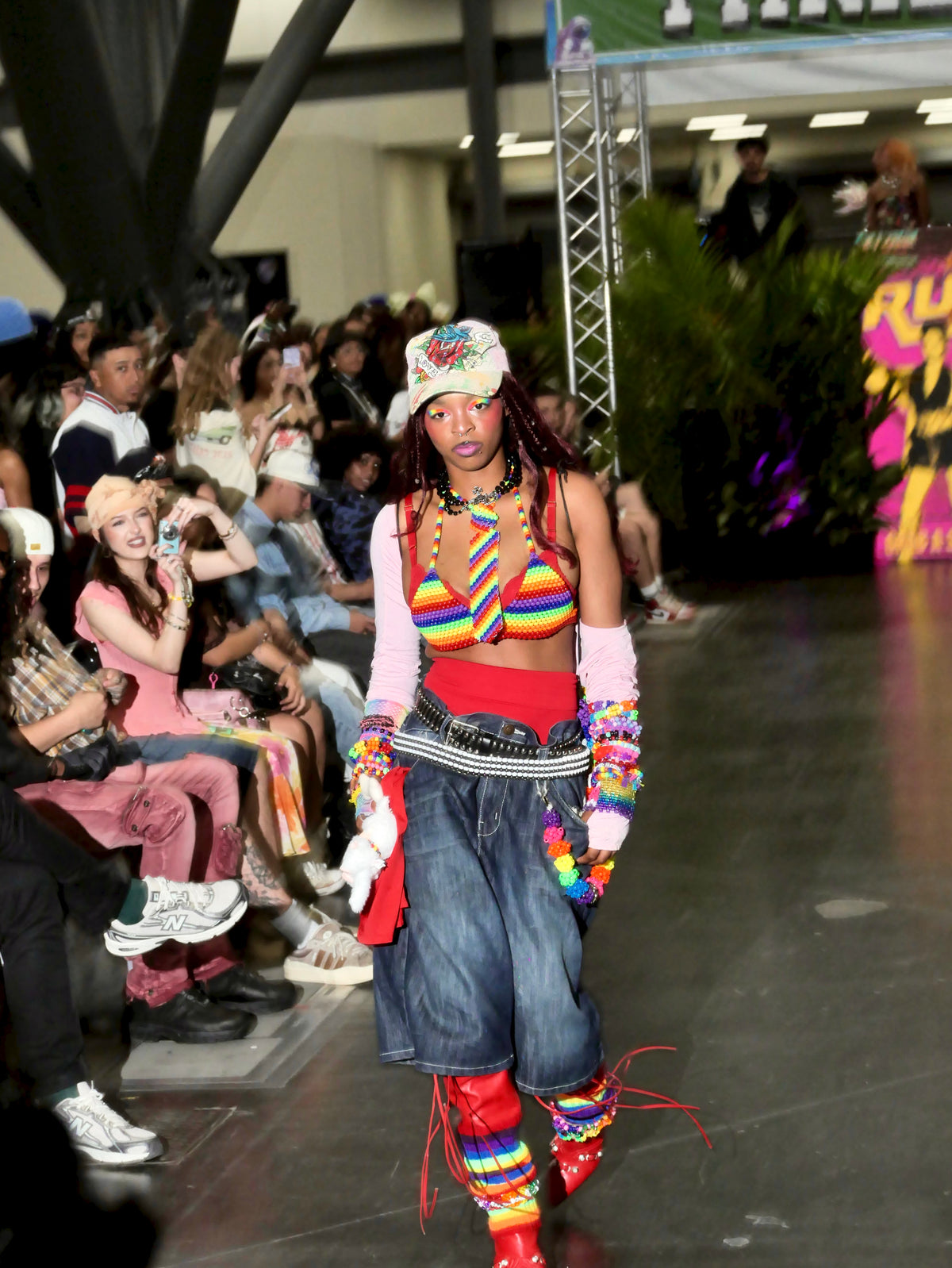Model walking on a fashion show runway wearing colorful outfit with audience in the background