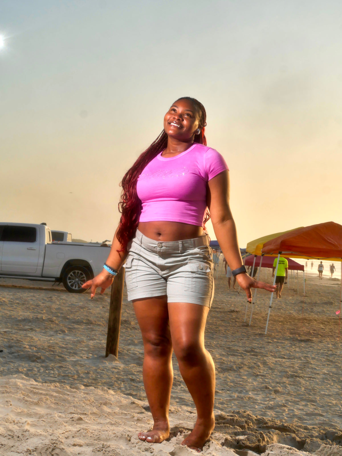 A woman standing on a sandy beach wearing a bright pink short-sleeve crop top and light gray denim shorts, with a pickup truck and beach umbrellas in the background.
