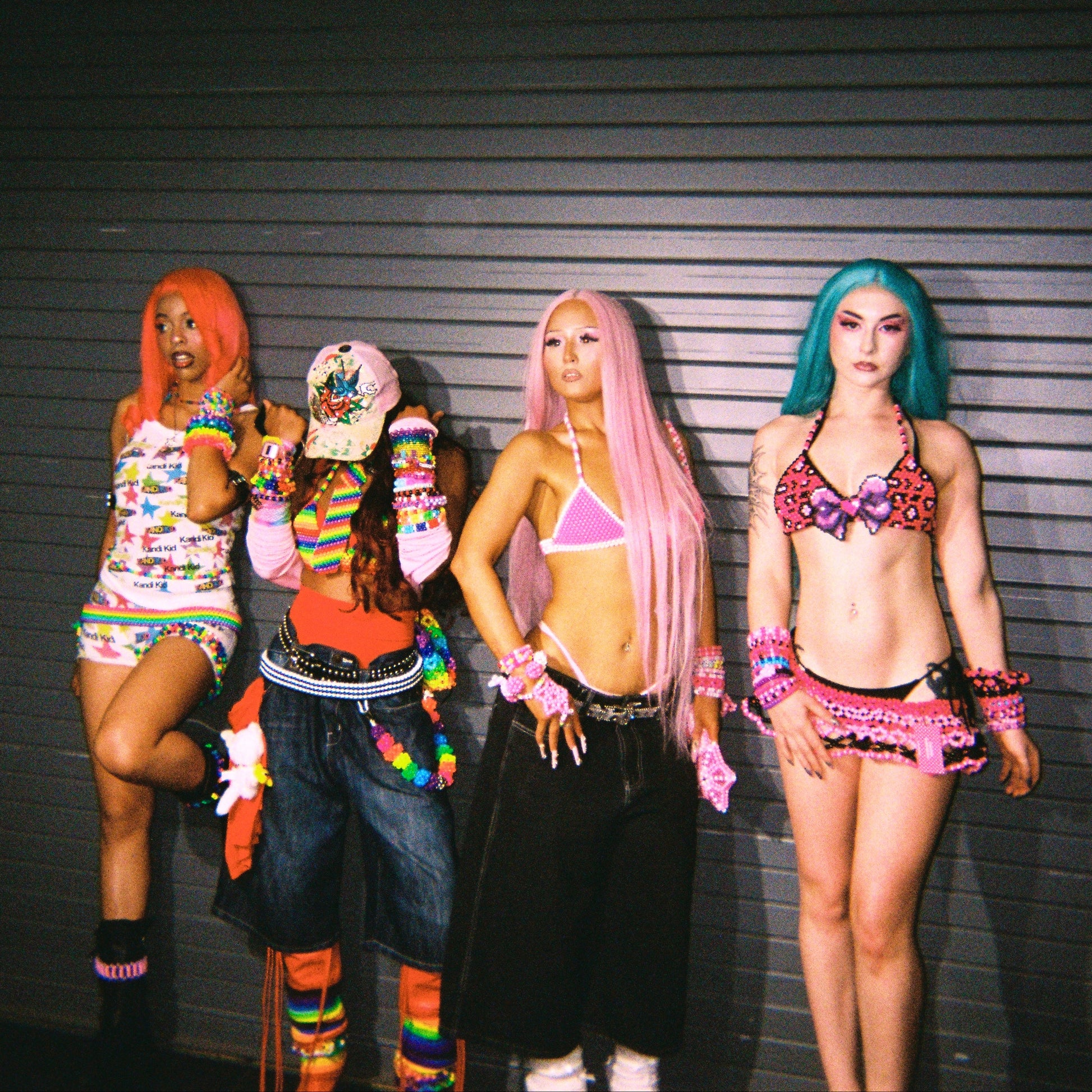 Four women posing in colorful, alternative-style outfits against a dark gray slatted wall.