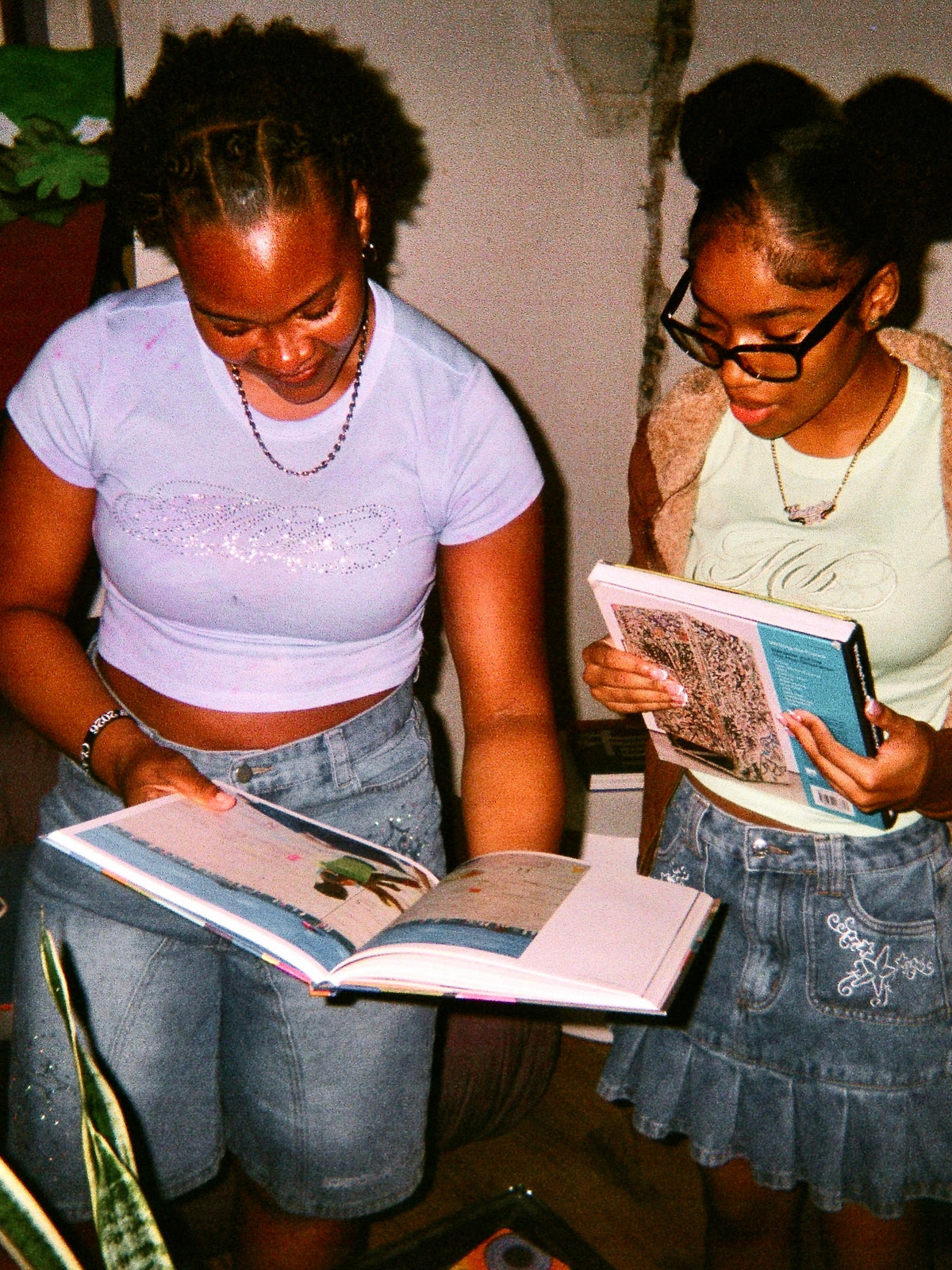 Two young women standing indoors and looking at open books. The woman on the left wears a light lavender crop top and denim shorts, and the woman on the right wears a pale green embroidered tee, denim skirt, and black-rimmed glasses.