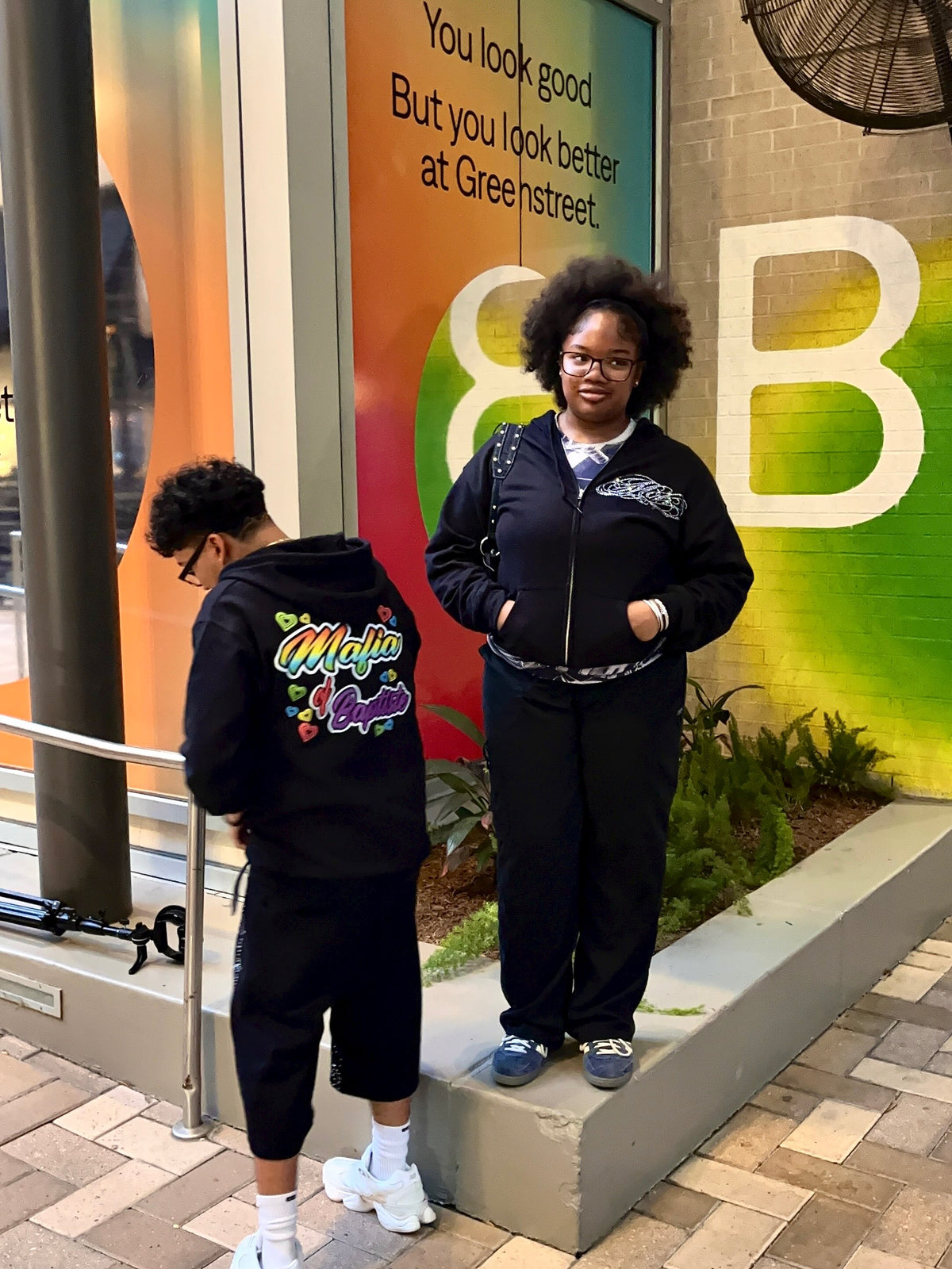 A woman and a man wearing matching black streetwear outfits, including black zip-up jackets and black sweatpants, standing outdoors in front of a colorful green and orange wall with the text 'You look good But you look better at Greensstreet.'.