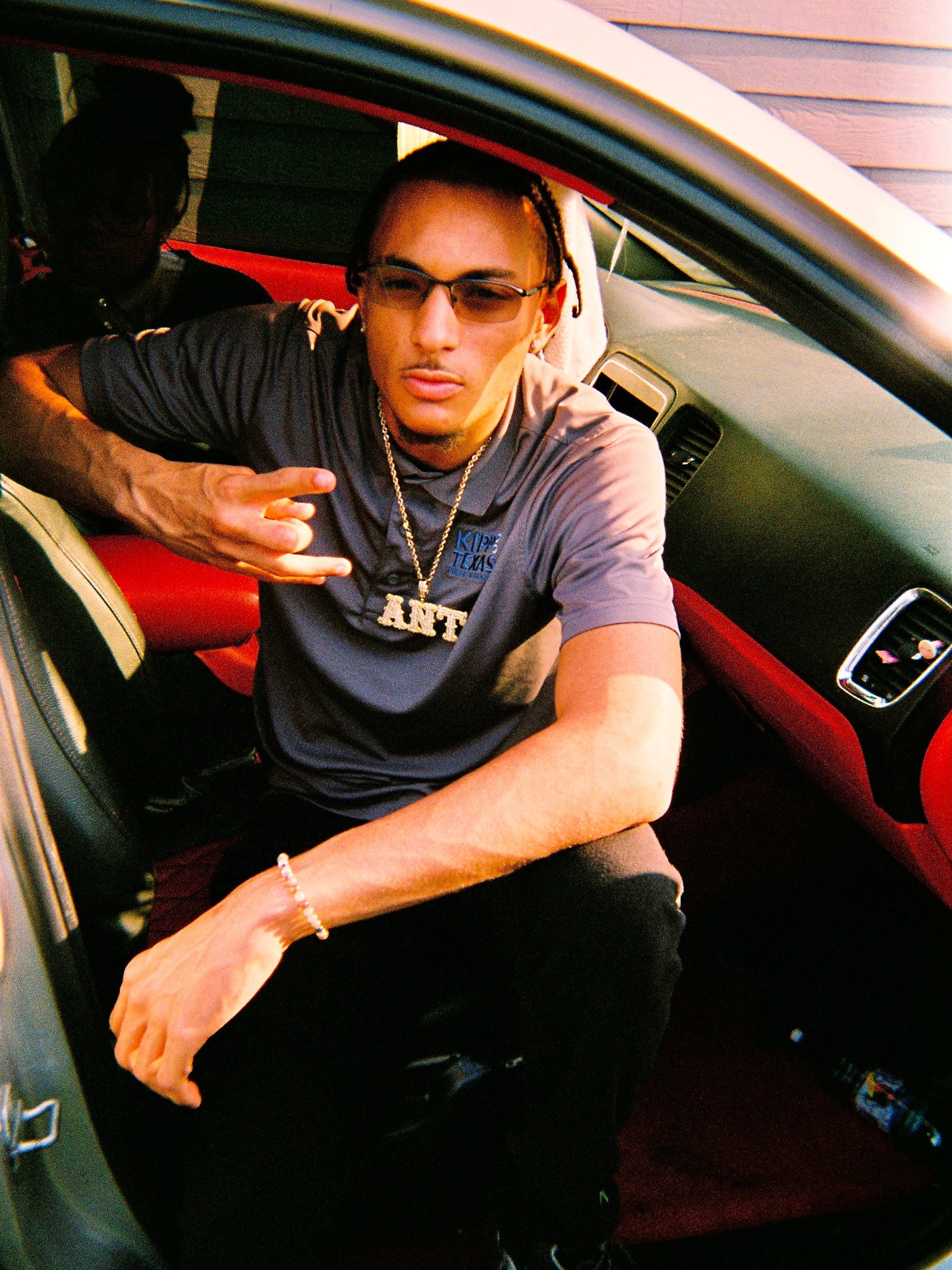 A man wearing a dark gray graphic t-shirt and sunglasses sits inside a car with a red and black interior, pointing toward the camera.