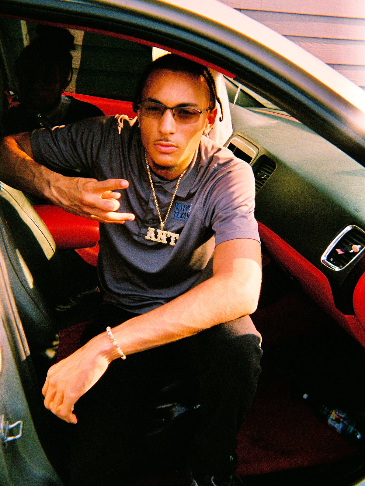 A man wearing a dark gray graphic t-shirt and sunglasses sits inside a car with a red and black interior, pointing toward the camera.